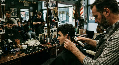 Close-up of a barber's hands using metal shears to precision cut a client's dampened hair in a dimly lit shop.