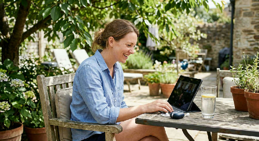 A man smiles during a video call on a tablet while sitting on a sunny patio with a glass of water nearby.