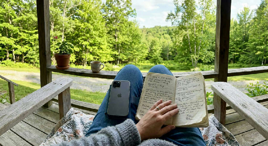 First-person view of a handwritten journal and a closed phone on a person's lap, looking out at a sunlit green yard.