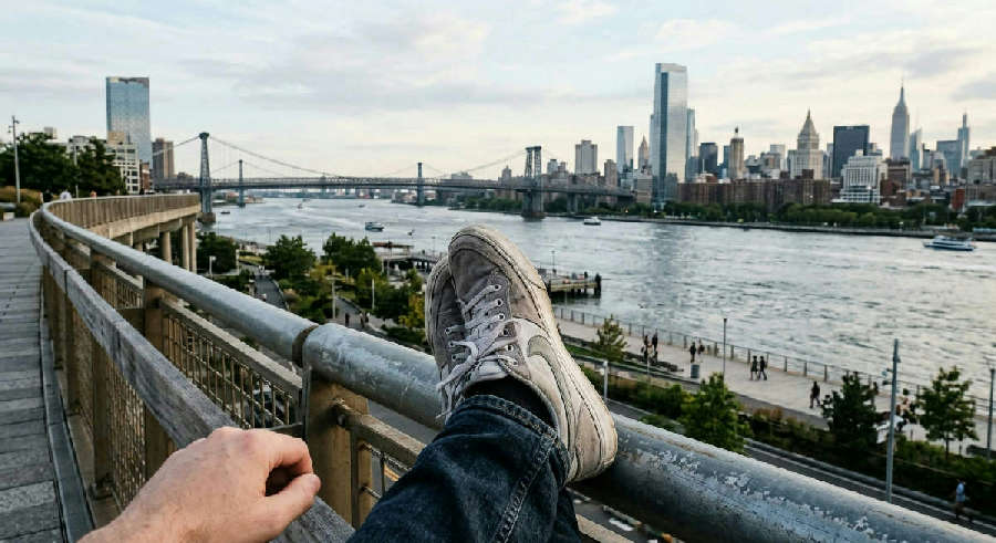 First-person view of feet in sneakers resting against a metal railing overlooking a calm river and distant city skyline.