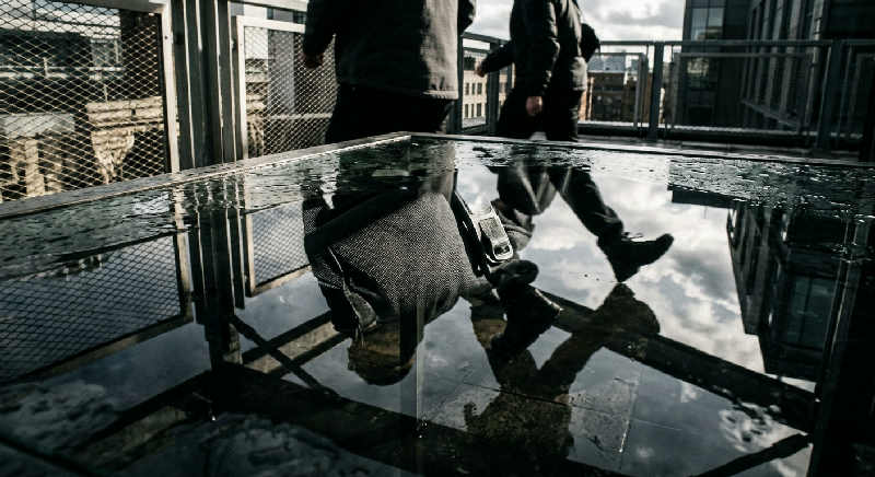 Close-up reflection on a dark glass surface showing a high-end black suit jacket and watch on a city terrace.